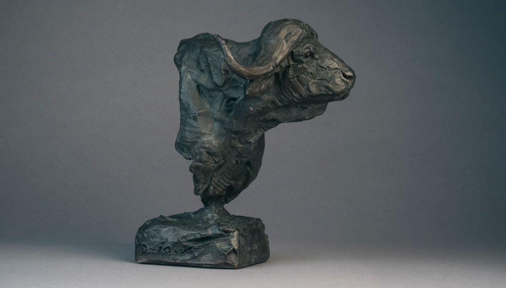 Bronze sculpture of a buffalo head with textured details on a rectangular base against a gray background.