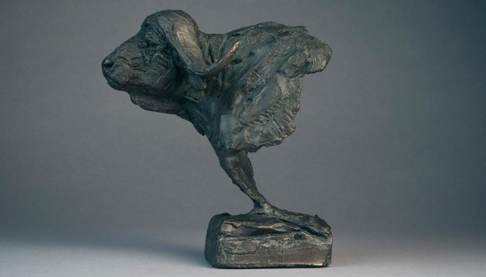 Bronze sculpture of a buffalo head on a rectangular base against a gray background.