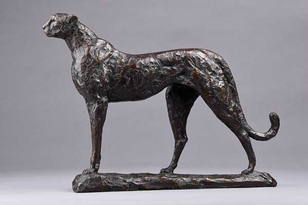 Side view of a textured bronze sculpture of a standing cheetah with a curled tail on a base.