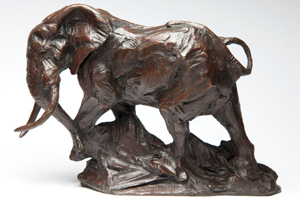 Bronze sculpture of an elephant standing on a textured base with its trunk lifted and tusks visible.