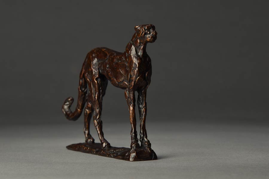 Bronze sculpture of a standing cheetah on a gray surface with a dark background.