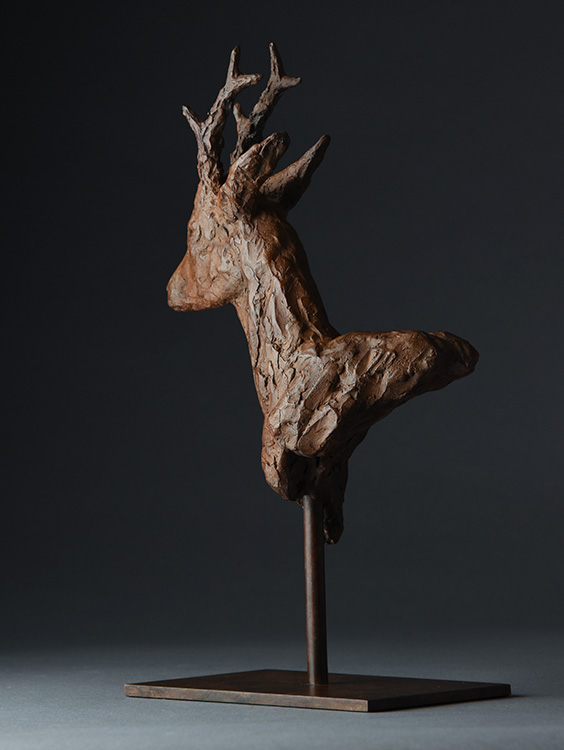 Bronze sculpture of a roe deer head with antlers on a rectangular base against dark background.