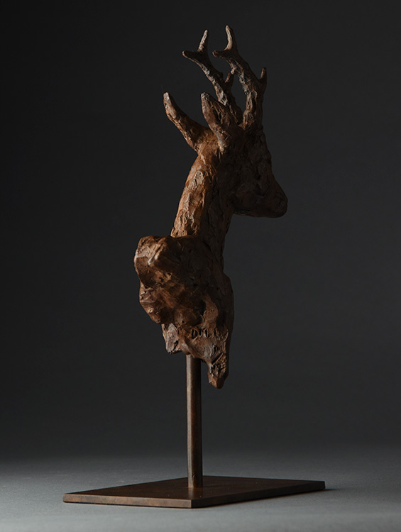 Side view of a textured bronze sculpture of a deer's head with antlers on a metal stand.