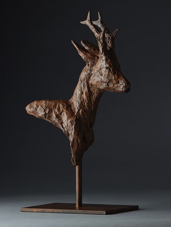 Brown textured sculpture of a deer's head and neck on a metal stand against dark background.
