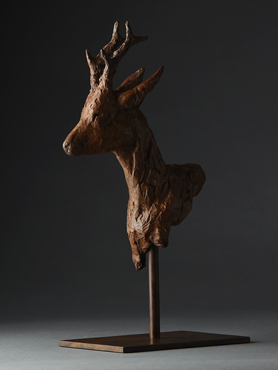 Bronze sculpture of a roe deer head with antlers on a vertical stand and flat base.