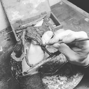 Hands sculpting a detailed lizard model using a small brush and clay on a workbench.